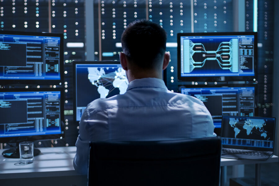 GRUPO HRO: System Security Specialist Working at System Control Center. Room is Full of Screens Displaying Various Information.